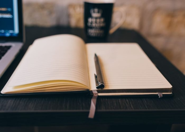 Open notebook with pen on a wooden desk, laptop, and a mug in a cozy setting.