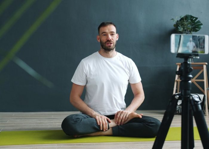A man in white T-shirt filming an online yoga session indoors using a smartphone on a tripod.